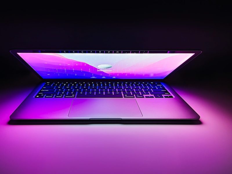 Ein halb geöffneter Apple Mac mit lila Licht vor schwarzen Hintergrund.