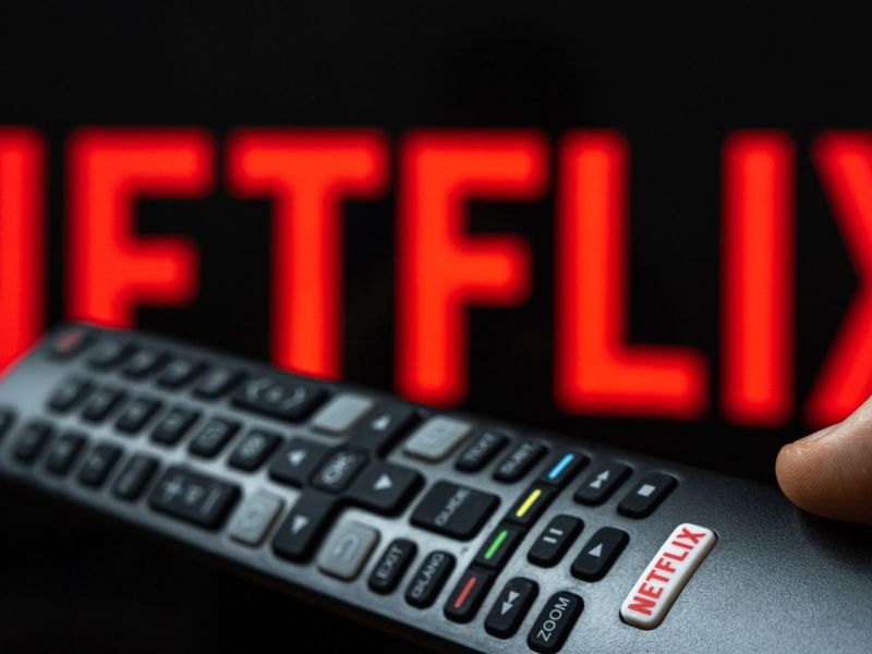 Netflix-Logo auf einem Fernseher, im Vordergrund Hand mit einer Fernbedienung.
