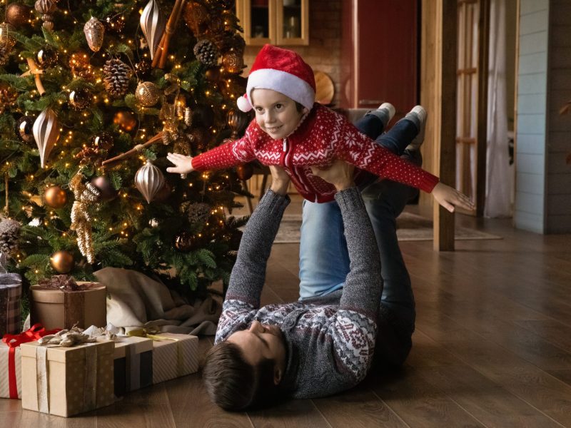 Mann spielt vor einem Weihnachtsbaum mit einem Kind Flugzeug