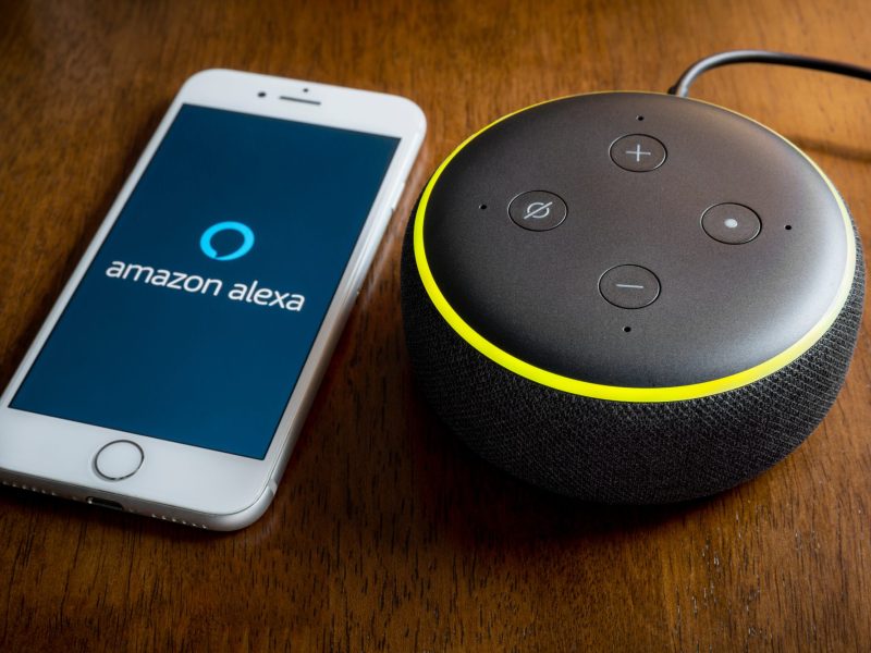 Amazon Echo liegt neben einem Smartphone, das die Alexa App anzeigt.