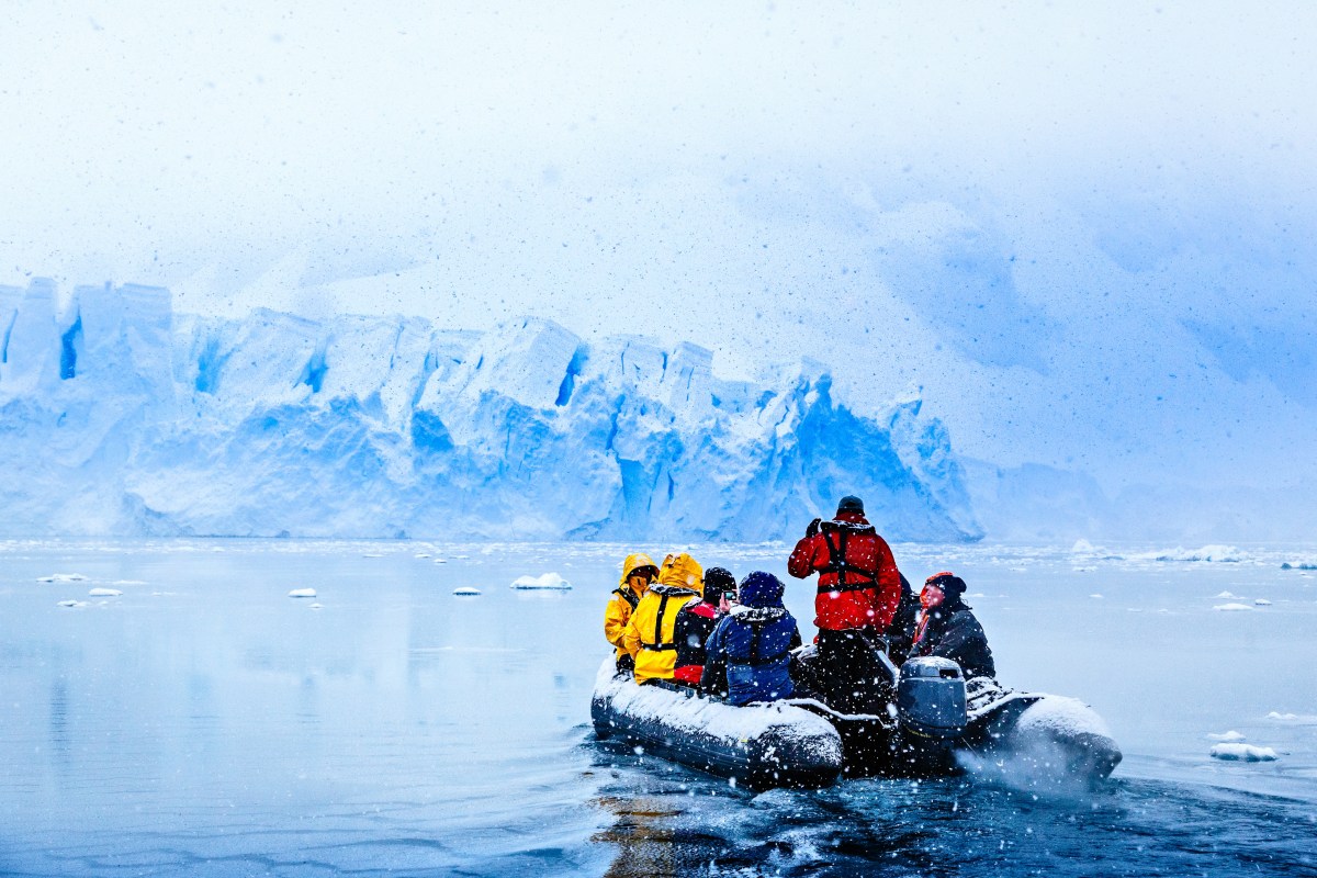Schneefall über einem Boot mit Touristen, die auf die riesige blaue Gletscherwand im Hintergrund zufahren
