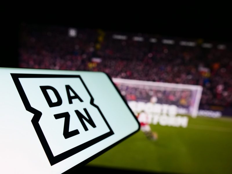 Die App von DAZN auf einem Smartphone; im Hintergrund steht ein Fußballtor.