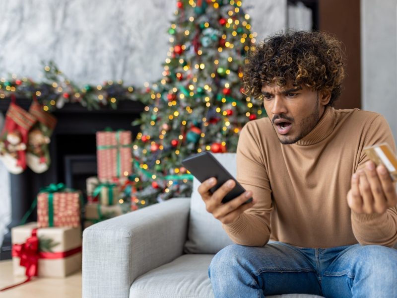 Ein Mann starrt an Weihnachten erbost auf sein Handy und seine Kreditkarte.