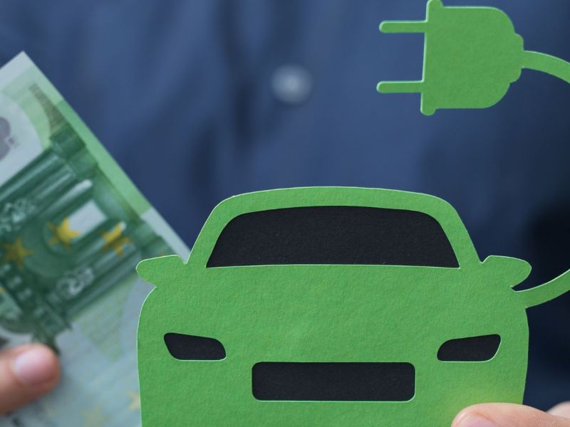 Peson hält Banknoten und ein Elektroauto aus Papier in Händen.