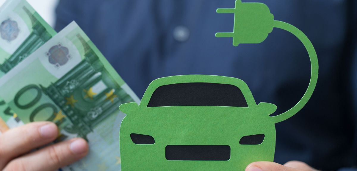 Peson hält Banknoten und ein Elektroauto aus Papier in Händen.