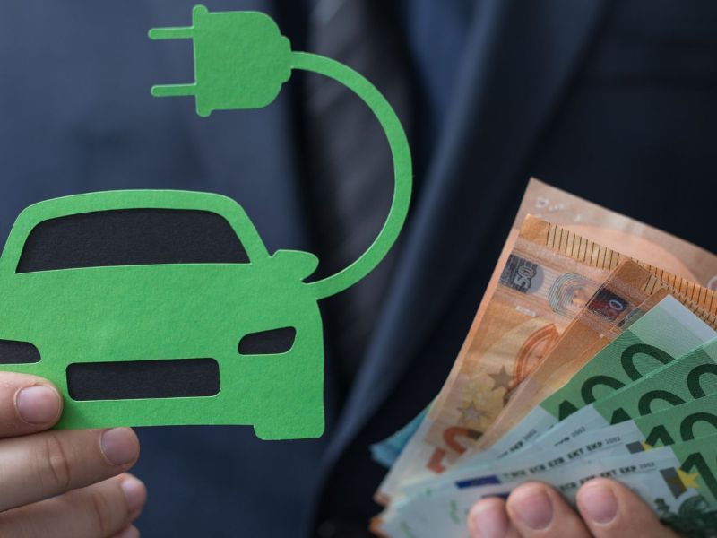 Person hält ein Elektroauto aus Papier sowie Banknoten in Händen.