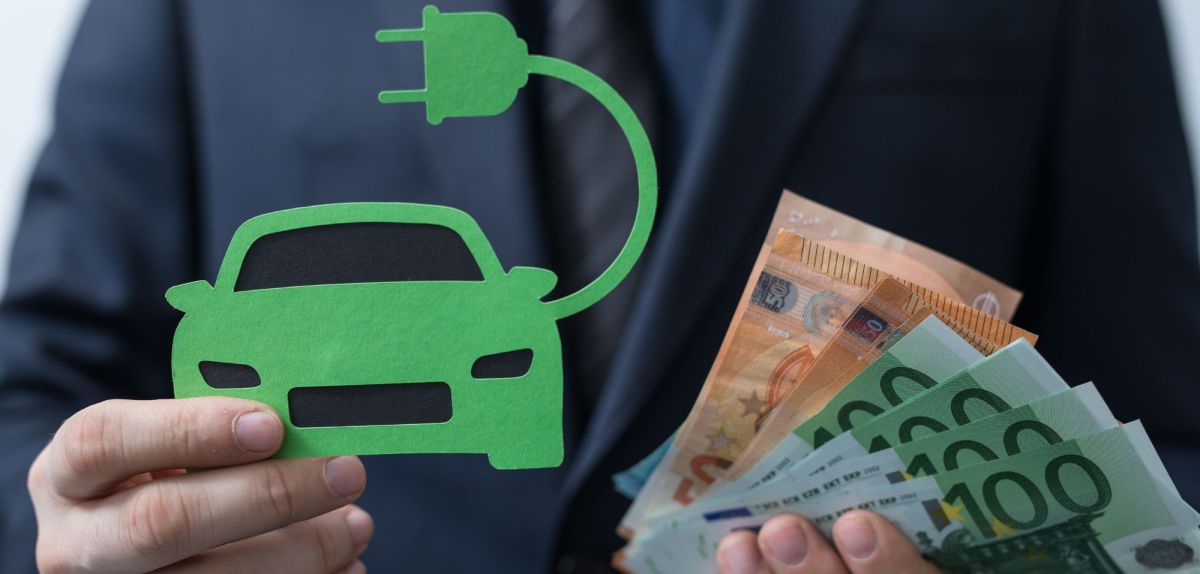 Person hält ein Elektroauto aus Papier sowie Banknoten in Händen.