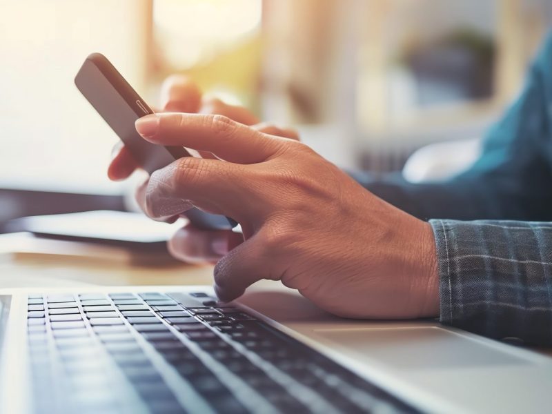 Person mit Smartphone vor einem Laptop