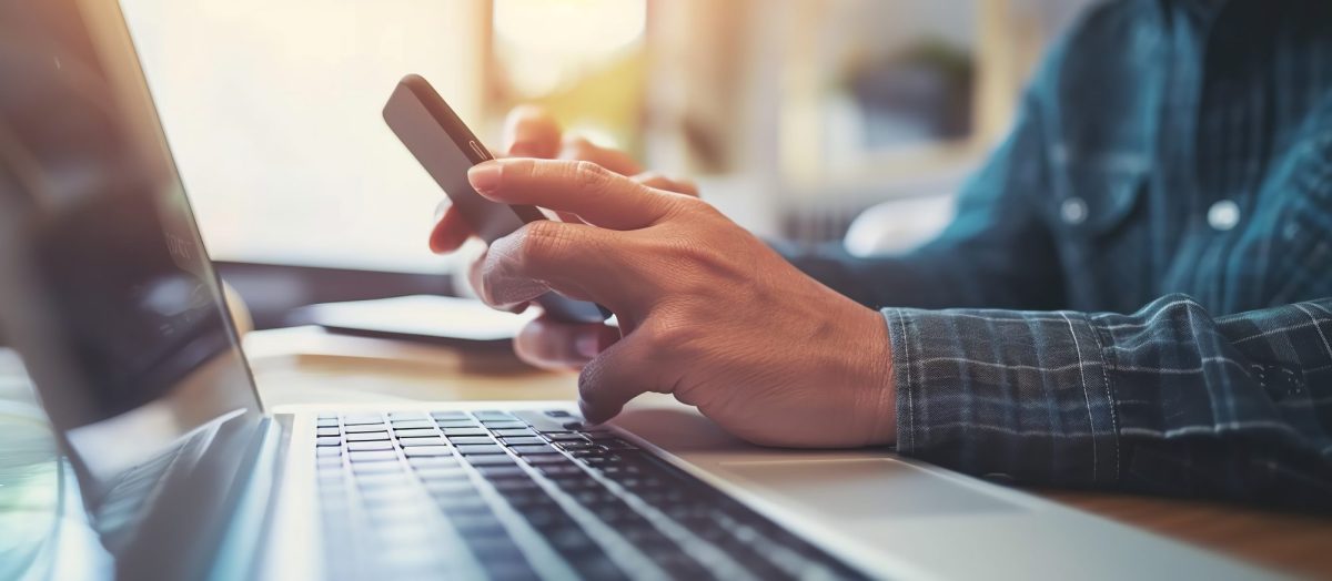 Person mit Smartphone vor einem Laptop