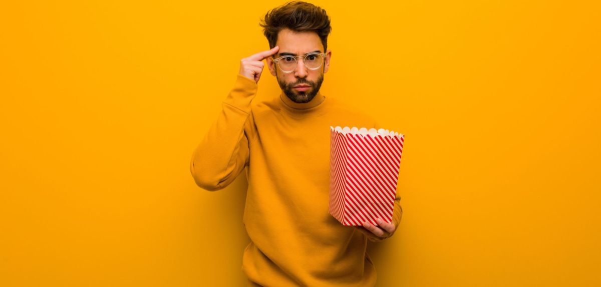 Mann hält eine Tüte Popcorn in der Händ und zeigt auf seinen Kopf.