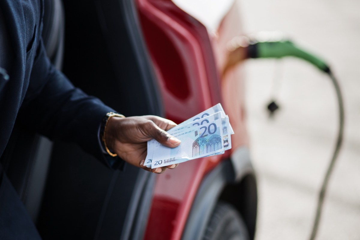 Mann hält Geldscheine vor einem Elektroauto in der Hand