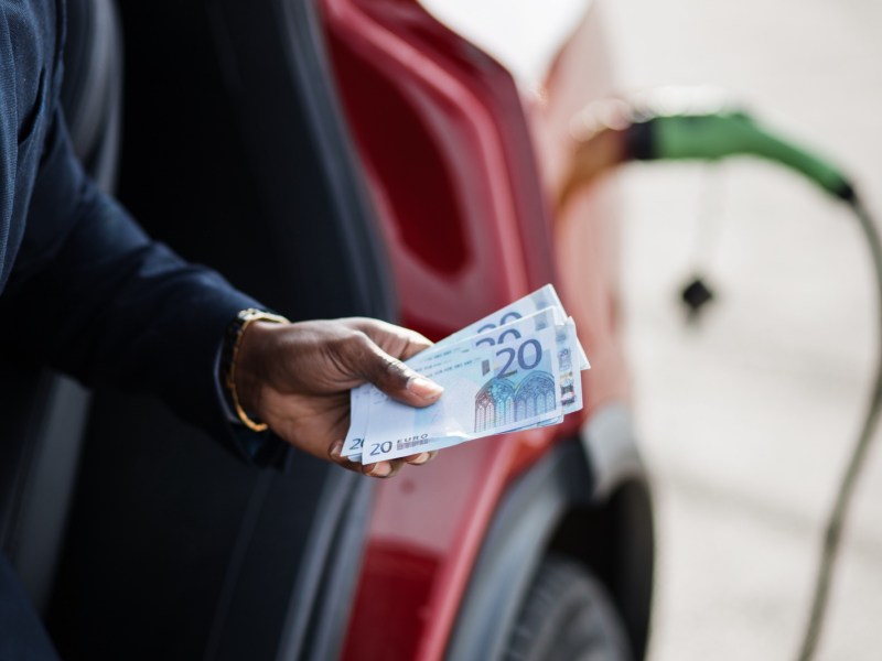 Mann hält Geldscheine vor einem Elektroauto in der Hand