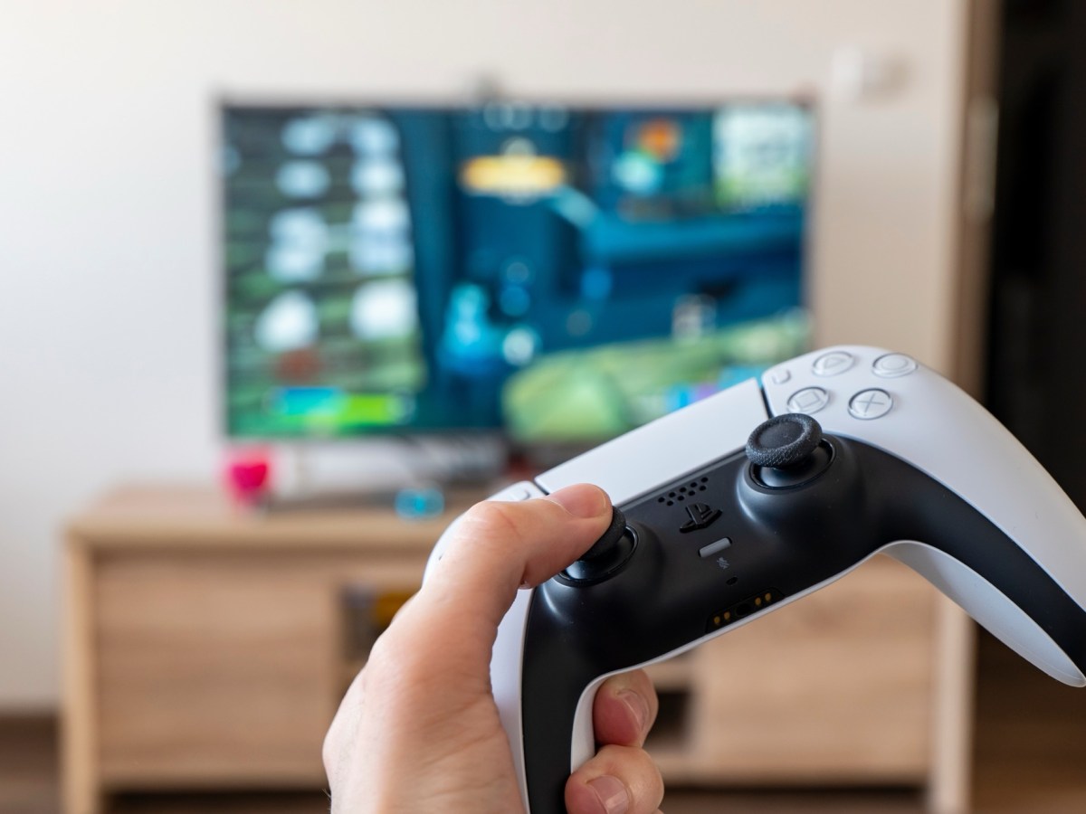 Mann hält einen PS5-Controller vor einen Fernseher.