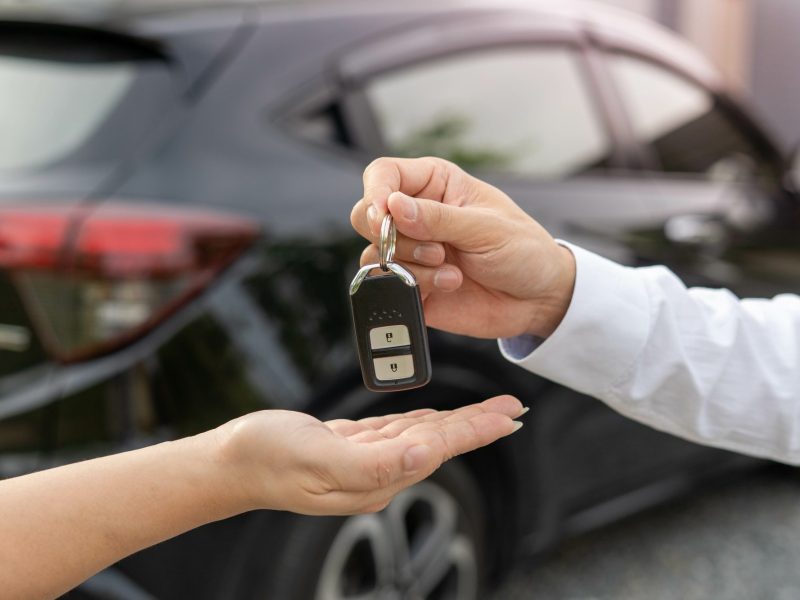 Person übergibt einer anderen Person einen Autoschlüssel. Im Hintergrund steht ein Auto.