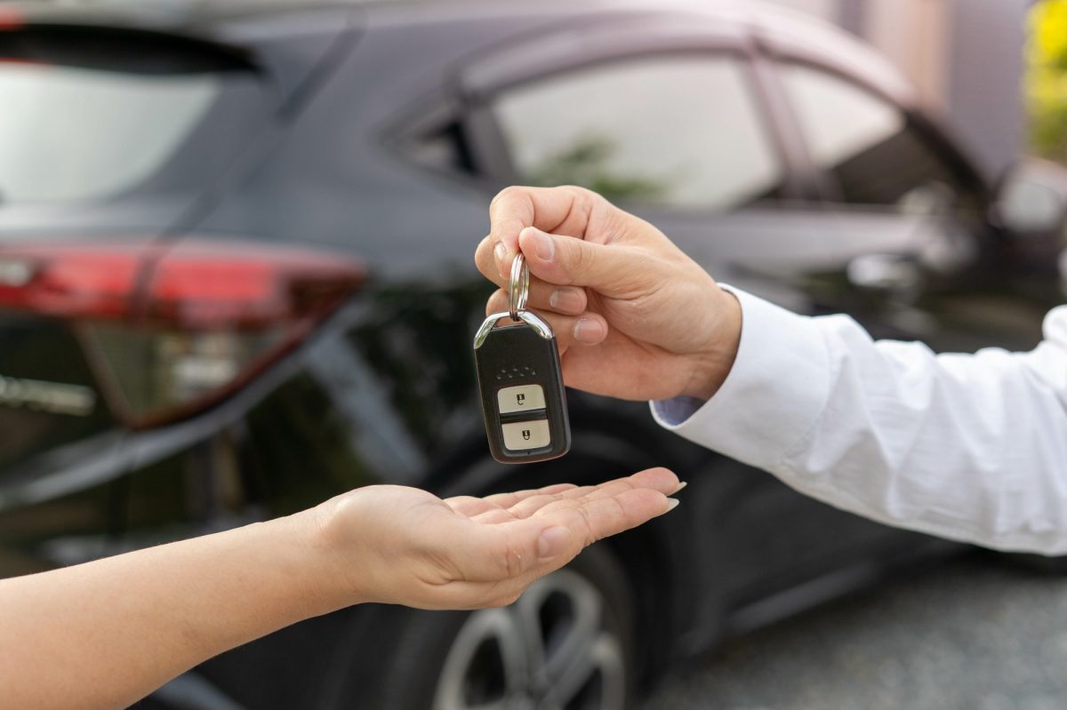Person übergibt einer anderen Person einen Autoschlüssel. Im Hintergrund steht ein Auto.