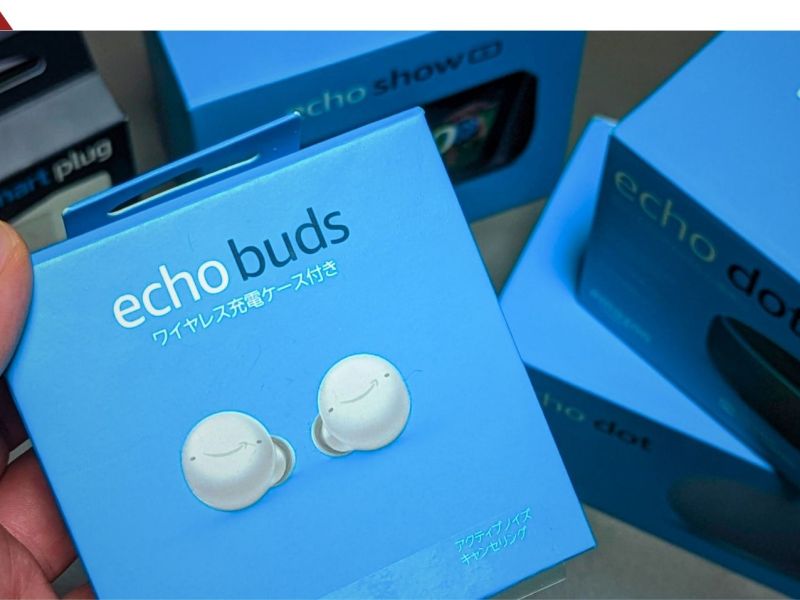 Eine Hand hält eine Packung Amazon Echo Buds. Dahinter liegen andere Produkte von Amazon.