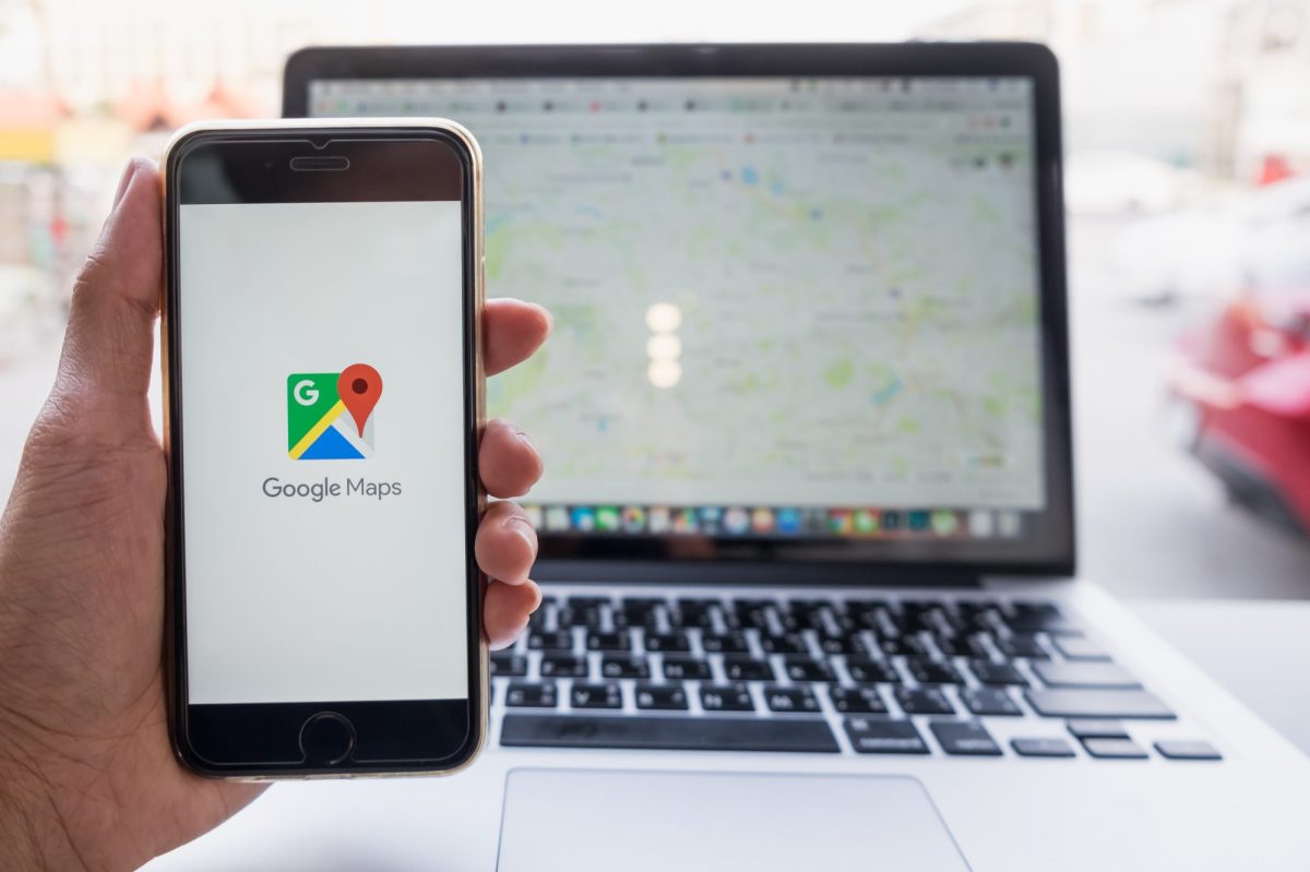 Eine Person hält ein Handy, auf dessen Display das Google Maps Logo angezeigt wird. Dahinter steht ein Laptop auf dem Google Maps geöffnet ist.