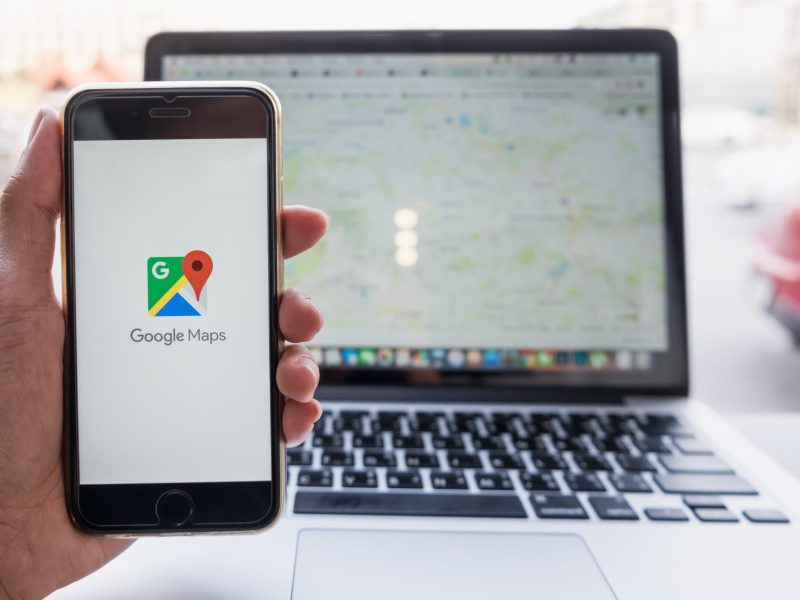 Eine Person hält ein Handy, auf dessen Display das Google Maps Logo angezeigt wird. Dahinter steht ein Laptop auf dem Google Maps geöffnet ist.