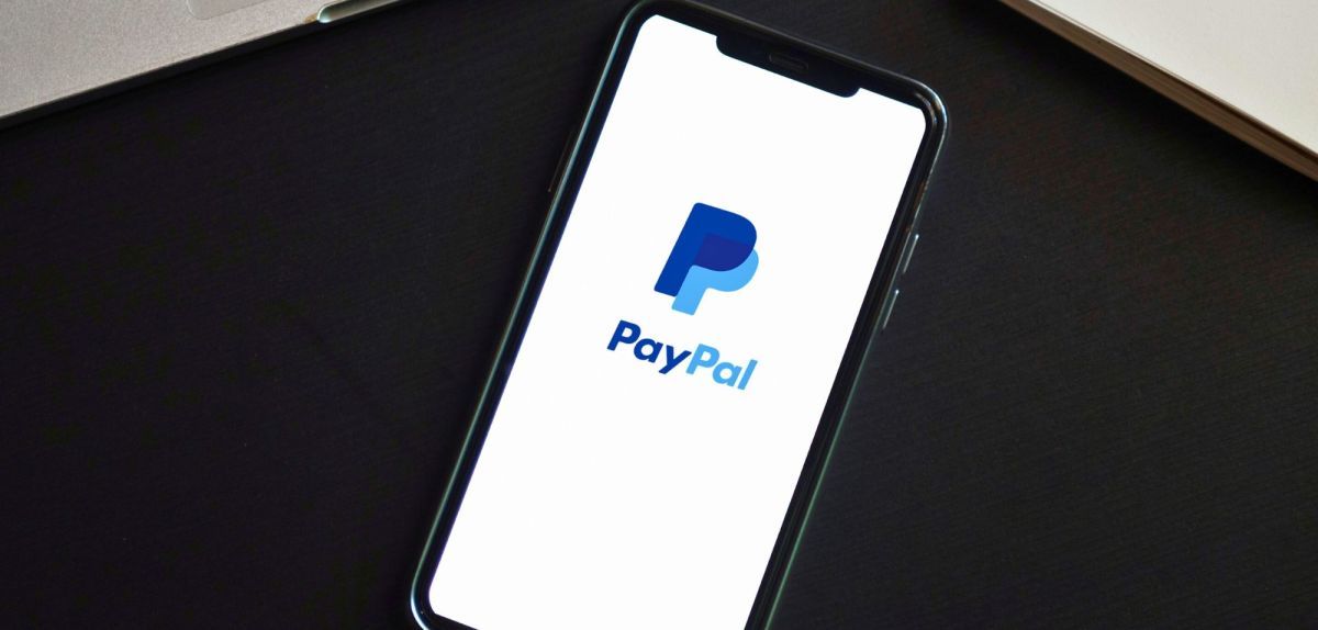 Smartphone mit PayPal-Logo zwischen Laptop und Notizbuch.