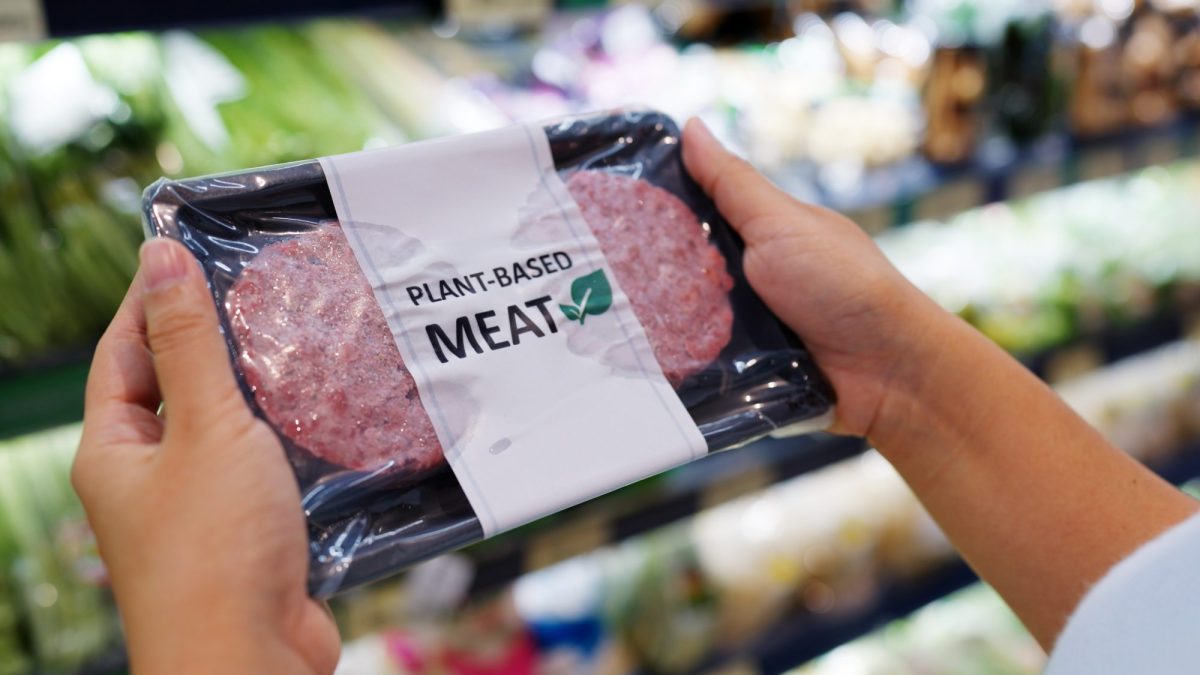 Person hält eine Packung eines pflanzlichen Fleischersatzproduktes vor einem Supermarktregal in der Hand.