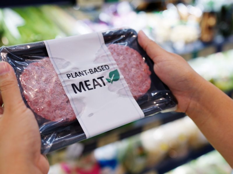 Person hält eine Packung eines pflanzlichen Fleischersatzproduktes vor einem Supermarktregal in der Hand.