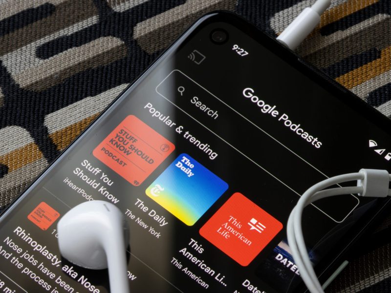 Auf einem Handy-Display ist die Google Podcasts-App geöffnet. Auf dem Handy liegen Kopfhörer.