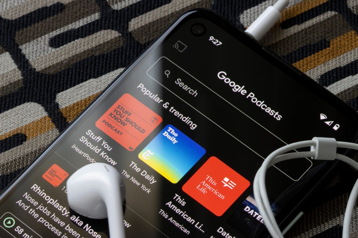 Auf einem Handy-Display ist die Google Podcasts-App geöffnet. Auf dem Handy liegen Kopfhörer.