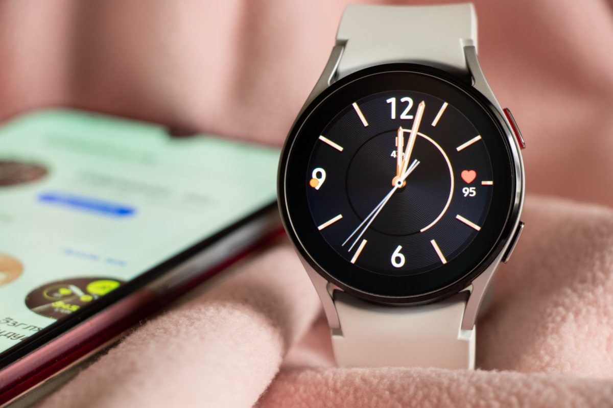 Samsung Galaxy Watch liegt auf einem rosafarbenen Untergrund. Im Hintergrund liegt ein Smartphone.