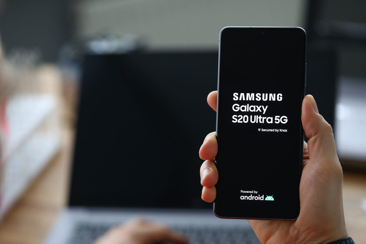 Eine Person hÃ¤lt ein Samsung Galaxy S20-handy in der Hand. Im Hintergrund ist ein aufgeklappter Laptop zu sehen.