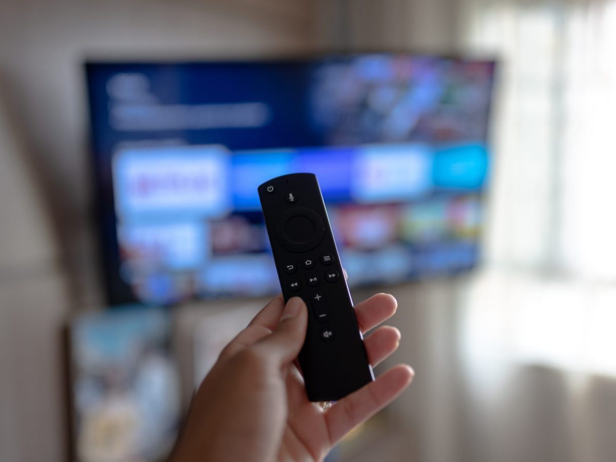 Person hält die Fernbedienung des Amazon Fire TV Sticks auf einen angeschalteten Fernseher gerichtet.