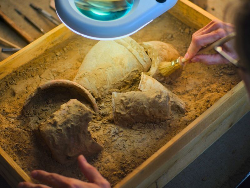 Nahaufnahme einer Archäologin, die unter einer Lampe mit einer Bürste das kulturelle Erbe einer untergegangenen Zivilisation aus dem Sand entfernt