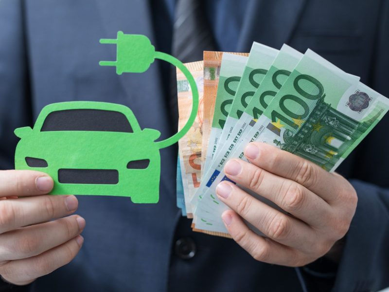 Eine Person hÃ¤lt in der einen Hand eine Zeichnung eines Elektroautos und in der anderen Hand mehrere Euroscheine.