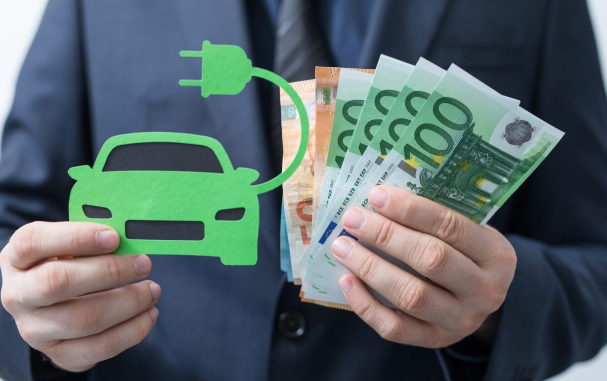 Eine Person hält in der einen Hand eine Zeichnung eines Elektroautos und in der anderen Hand mehrere Euroscheine.