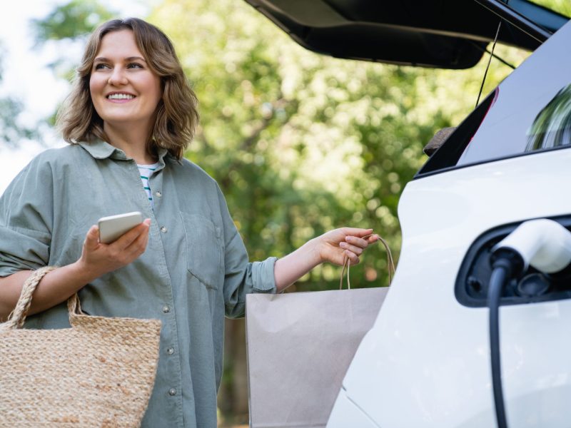 Frau lÃ¤dt EinkÃ¤ufe in den Kofferraum, wÃ¤hrend ihr Elektroauto geladen wird.