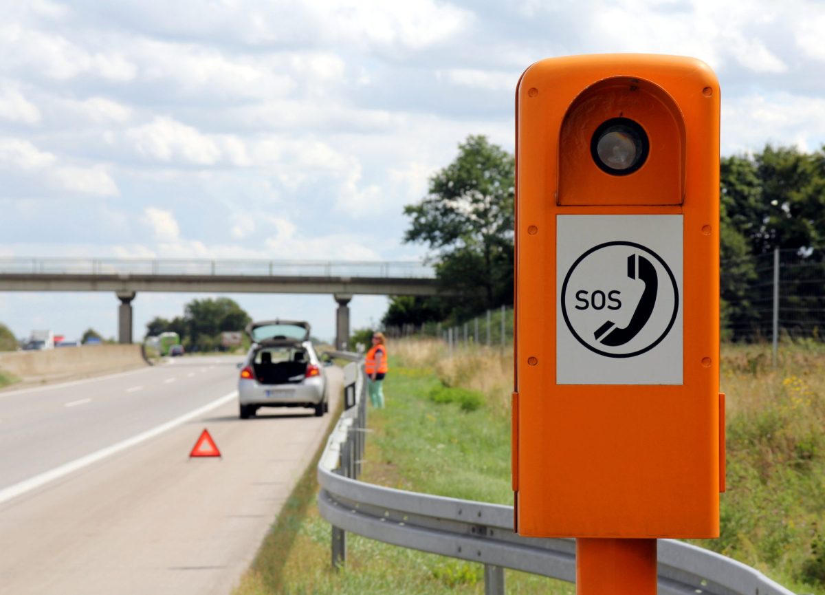 SOS-Säule an einer Autobahn. Im Hintergrund ist ein Auto zu sehen, das hinter einem Warndreieck auf dem Seitenstreifen steht.
