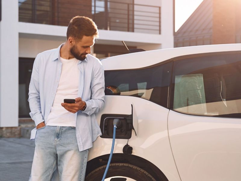 Aufladen des Fahrzeugs in der Nähe des Hauses. Hält Smartphone. Junge stilvolle Mann ist mit Elektroauto am Tag.