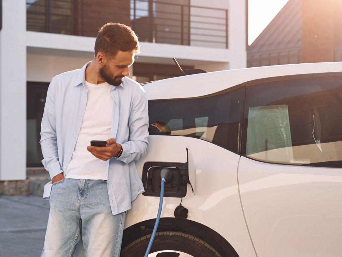 Aufladen des Fahrzeugs in der Nähe des Hauses. Hält Smartphone. Junge stilvolle Mann ist mit Elektroauto am Tag.