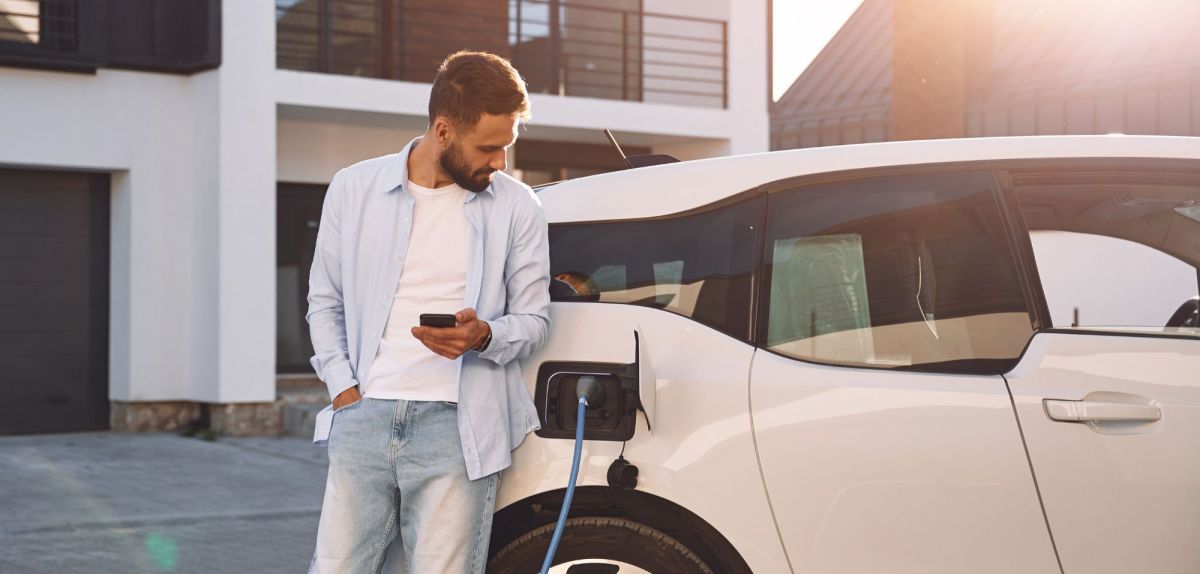 Aufladen des Fahrzeugs in der Nähe des Hauses. Hält Smartphone. Junge stilvolle Mann ist mit Elektroauto am Tag.