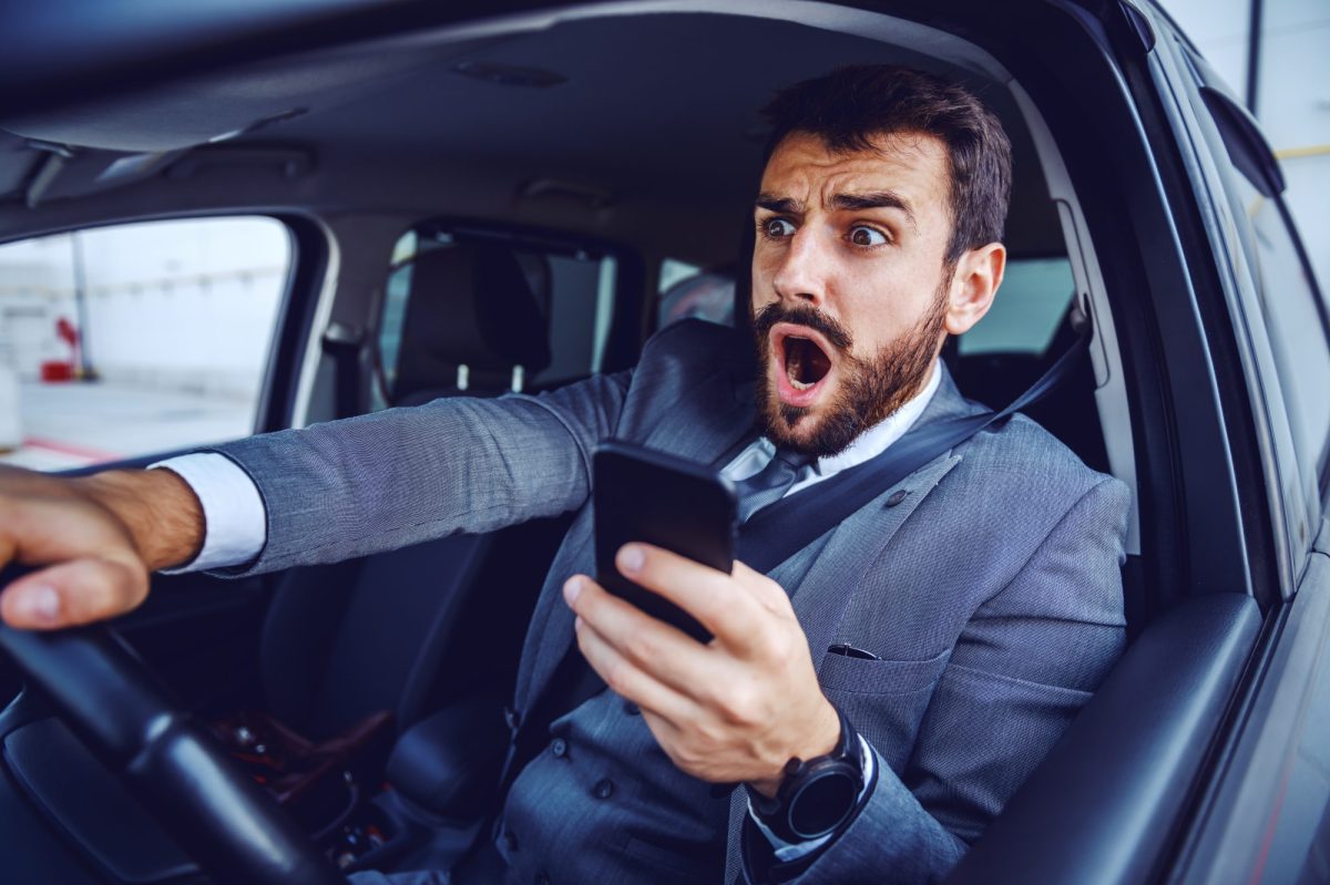 Mann am Steuer hält ein Handy in der Hand und macht ein erschrockenes Gesicht.