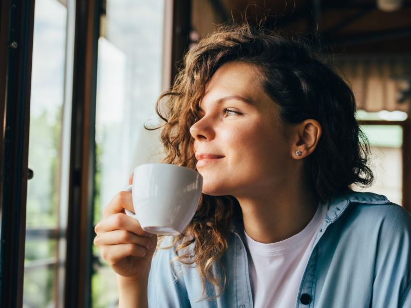 Frau mit einer weißen Tasse blickt aus einem Fenster