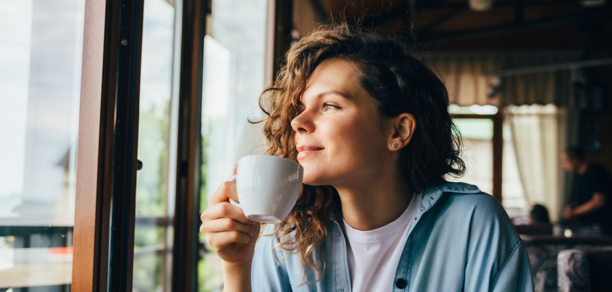 Frau mit einer weißen Tasse blickt aus einem Fenster