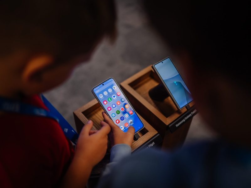 Zwei Personen stehen vor einem Samsung-Handy, auf dem mehrere Apps angezeigt werden.