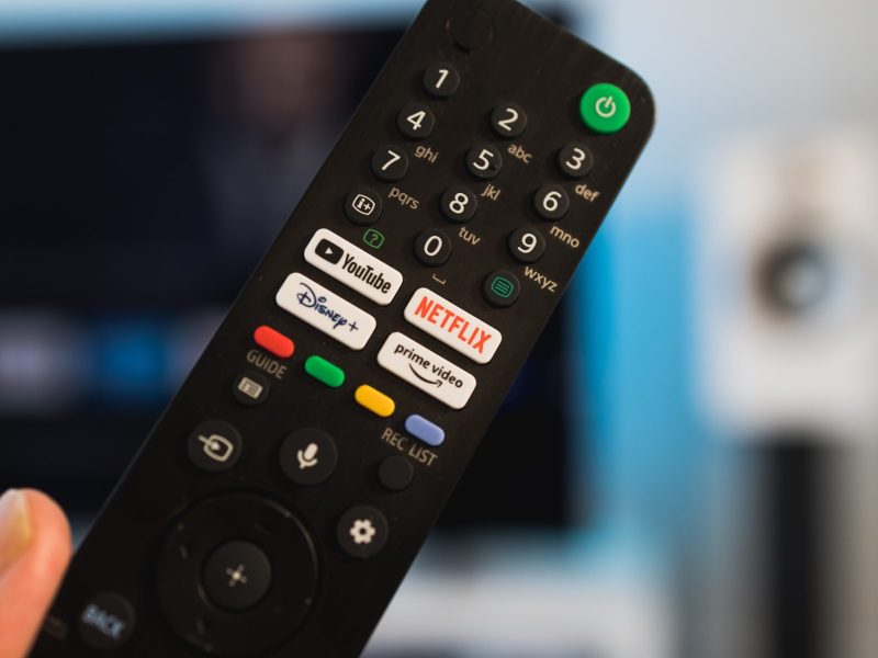 Person hält eine Fernbedienung. Im Hintergrund ist ein Fernseher zu sehen.