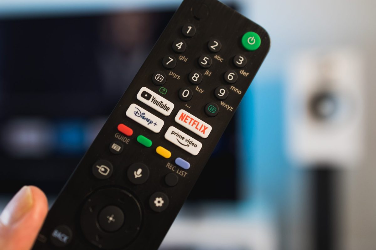 Person hält eine Fernbedienung. Im Hintergrund ist ein Fernseher zu sehen.