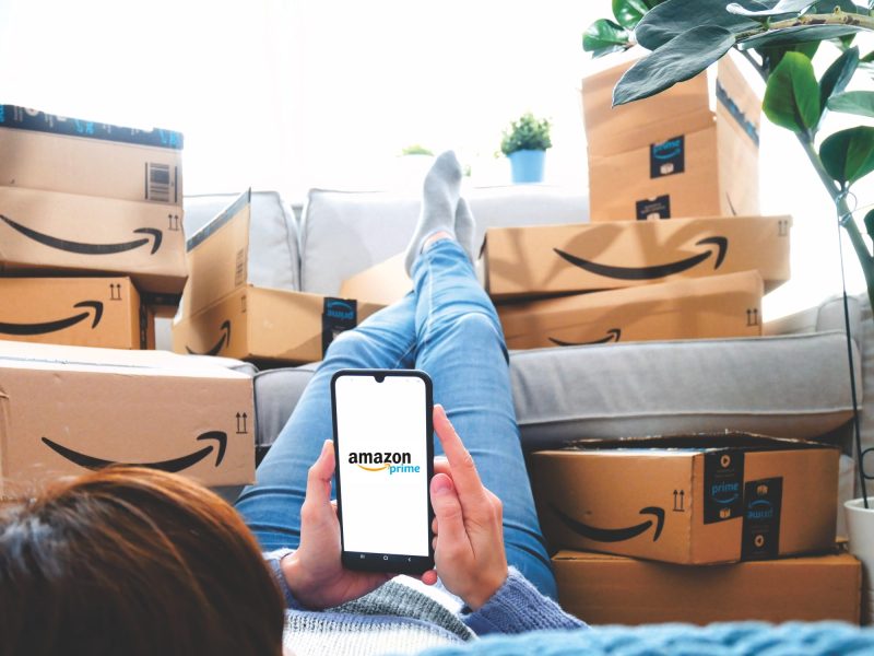 Person hält ein Handy in der Hand, auf dessen Display das Amazon Prime-Logo angezeigt. Daneben liegen mehrere Amazon-Pakete.