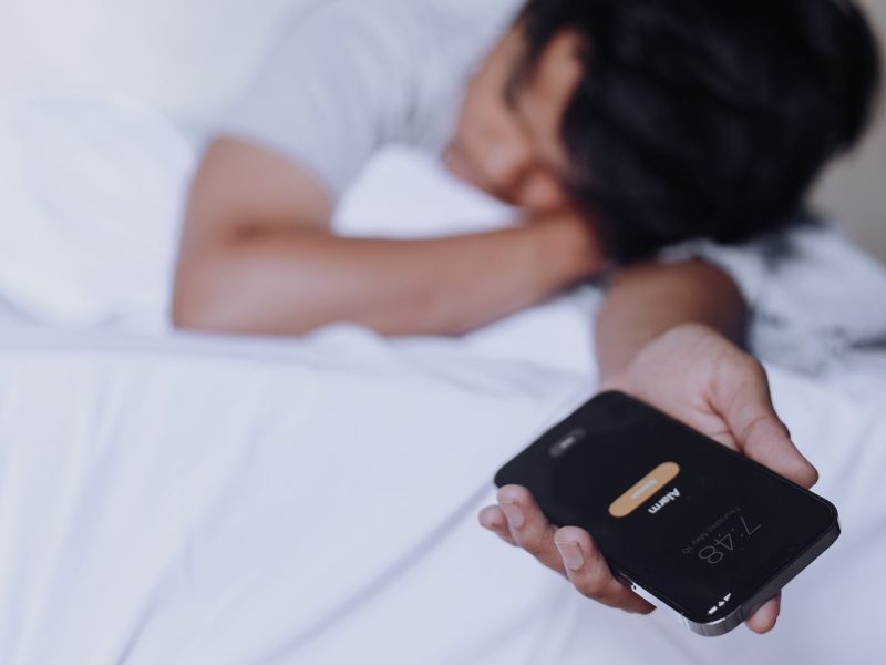 Person liegt im Bett und hält ein Handy in der Hand, auf dem ein Wecker klingelt.