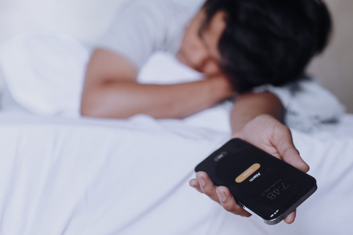 Person liegt im Bett und hält ein Handy in der Hand, auf dem ein Wecker klingelt.