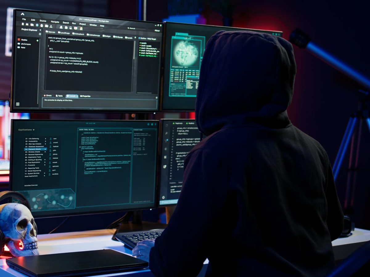Person mit Kapuze sitzt im Dunkeln vor mehreren Computer-Bildschirmen.