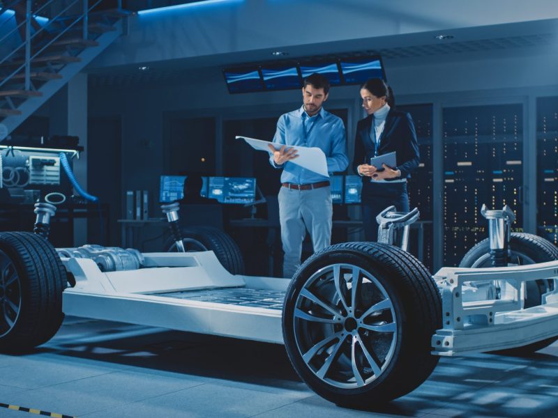 Personen sprechen über einen Bauplan, im Vordergrund steht eine Autokarosserie.
