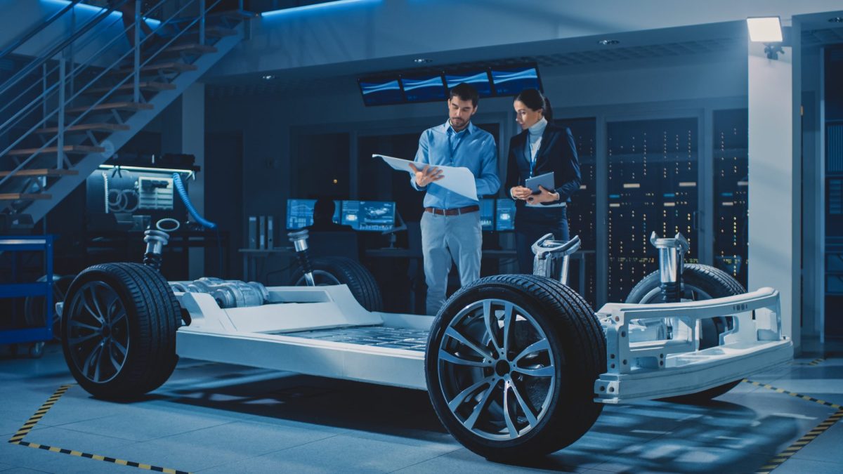 Personen sprechen über einen Bauplan, im Vordergrund steht eine Autokarosserie.
